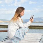 woman using smartphone while sitting of dock during daytime